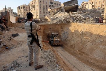 Un combatiente de la resistencia palestina observa una excavadora mientras excava en busca de cuerpos de prisioneros israelíes en Jan Yunis (AFP)